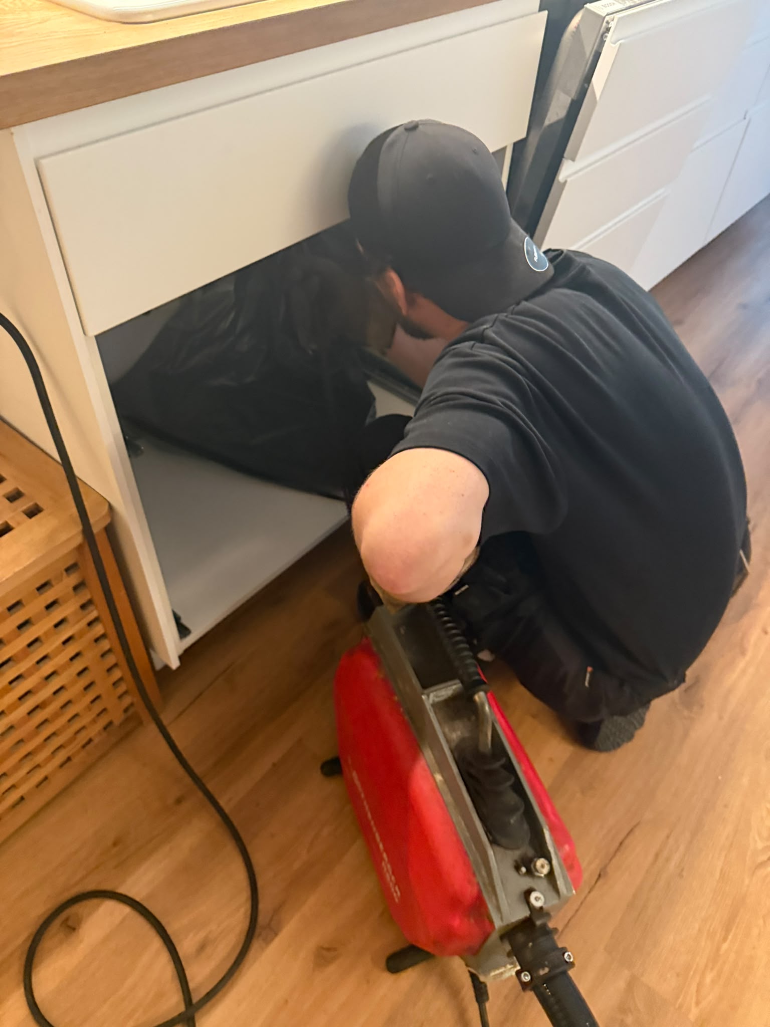 Monteur kniet vor einem geöffneten Küchenunterschrank und reinigt den Abfluss mit einer elektrischen Rohrreinigungsmaschine