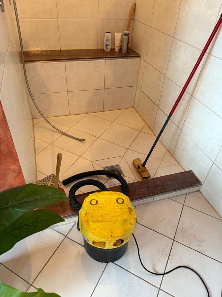 Geflieste Dusche mit geöffnetem Bodenablauf, davor Nasssauger, Saugglocke und Schrubber beim Freimachen des Abflusses