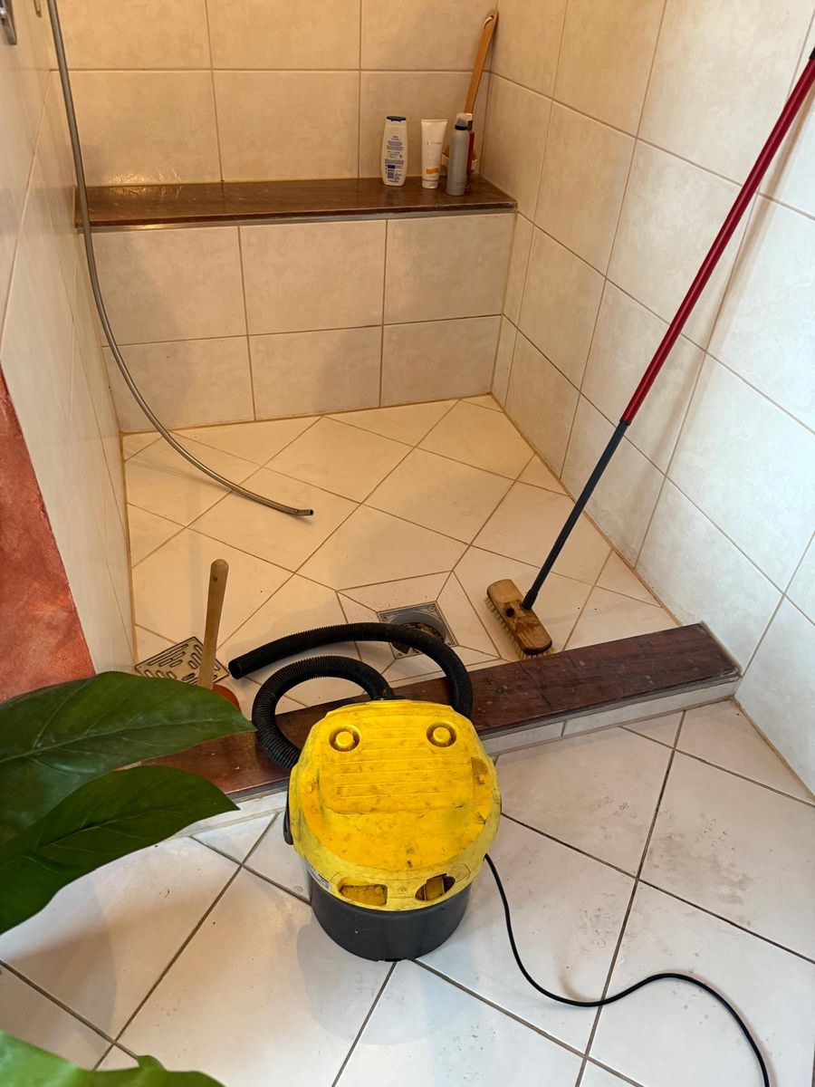 Geflieste Dusche mit geöffnetem Bodenablauf, davor Nasssauger, Saugglocke und Schrubber beim Freimachen des Abflusses
